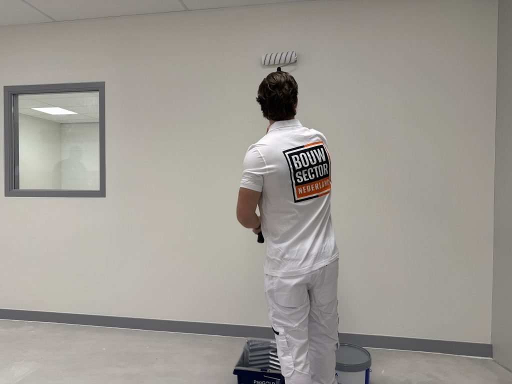 Schilder van Bouwsector Nederland zet een witte muur met de roller, met verfbak en emmer aan zijn voeten