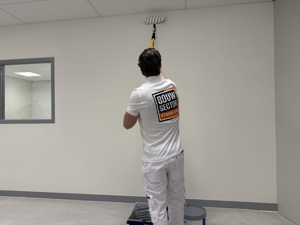 Schilder in wit werkshirt rolt met een roller op een verlengsteel de bovenkant van een lichte muur