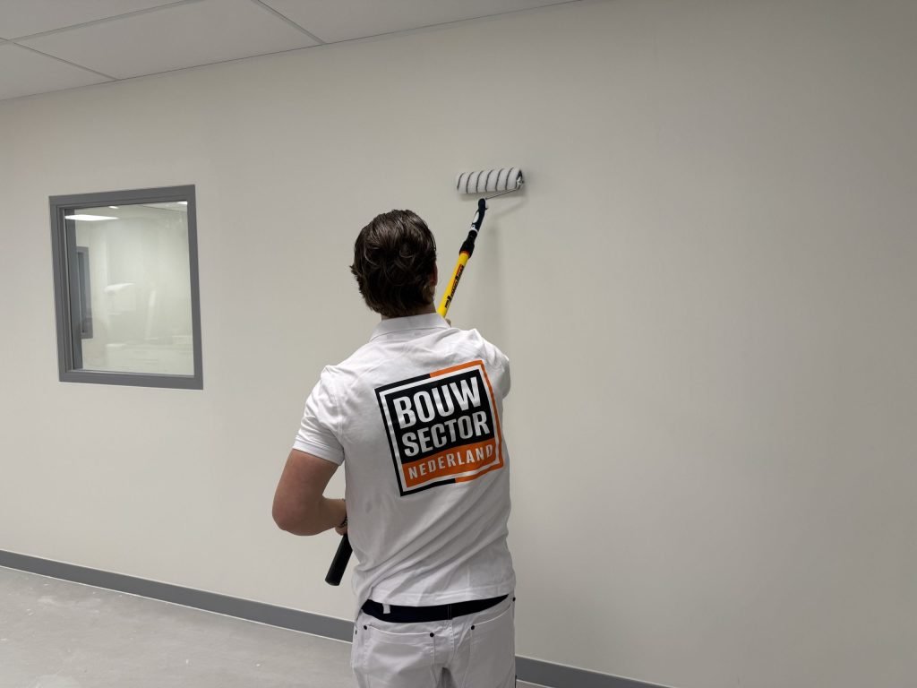 Schilder met Bouwsector-logo op zijn rug zet met de roller een baan verf op een lichte wand bij een binnenraam