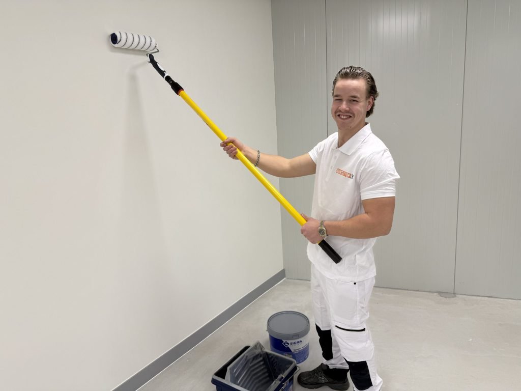 Lachende schilder in wit Bouwsector-shirt rolt een lichte muur met een roller aan een gele steel, naast een Sigma-emmer