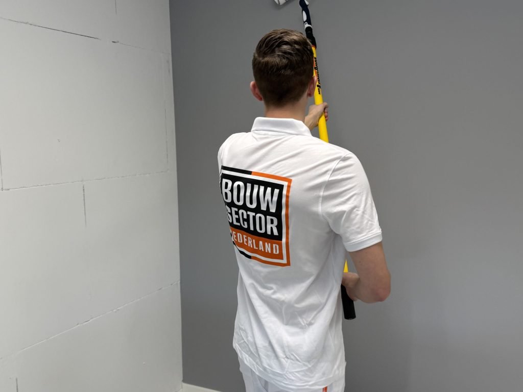Schilder rolt een hoge grijze wand met een lange steel, naast een ongeschilderde muur van blokken