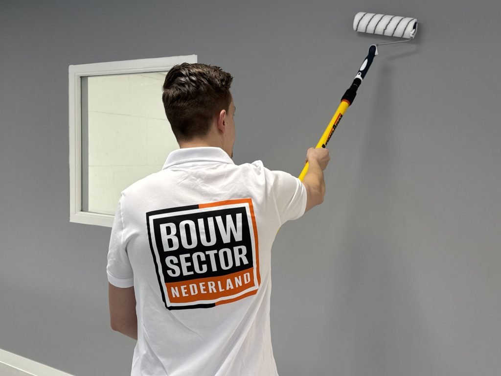 Schilder van Bouwsector Nederland rolt met een lange roller de bovenkant van een grijze wand