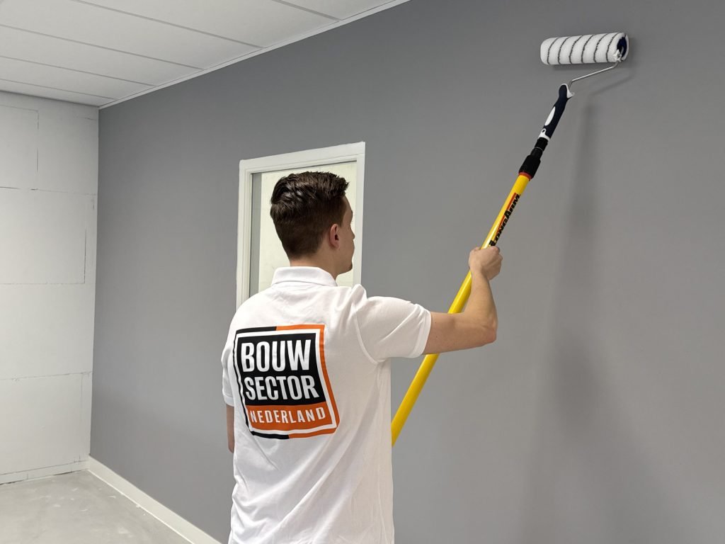 Schilder in wit poloshirt van Bouwsector Nederland rolt grijze muurverf op een wand naast een deur