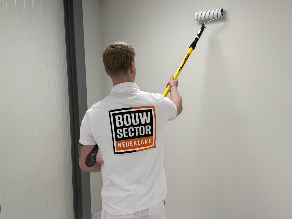 Schilder met het logo van Bouwsector Nederland rolt een lichtgrijze wand tot vlak onder het plafond