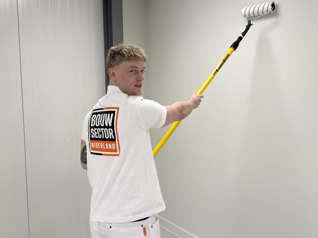 Schilder in wit werkshirt kijkt over zijn schouder terwijl hij een lichte gangmuur rolt
