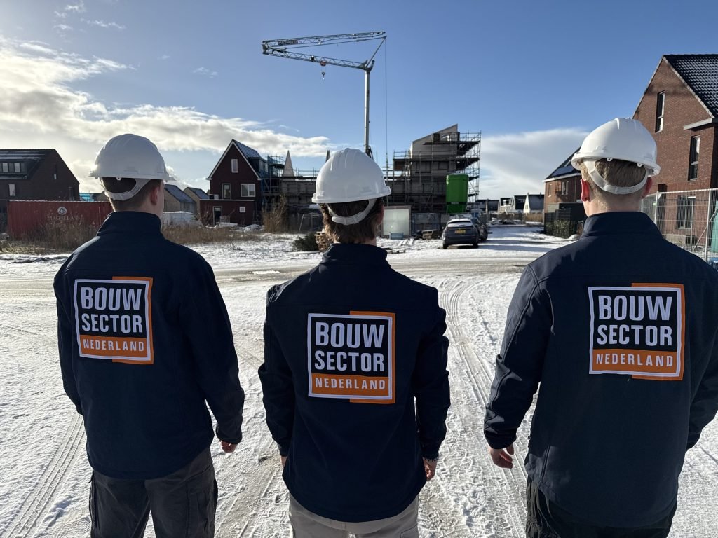 Drie vakmensen van Bouwsector Nederland met witte helm en donkerblauwe jas kijken naar nieuwbouwwoningen in de sneeuw