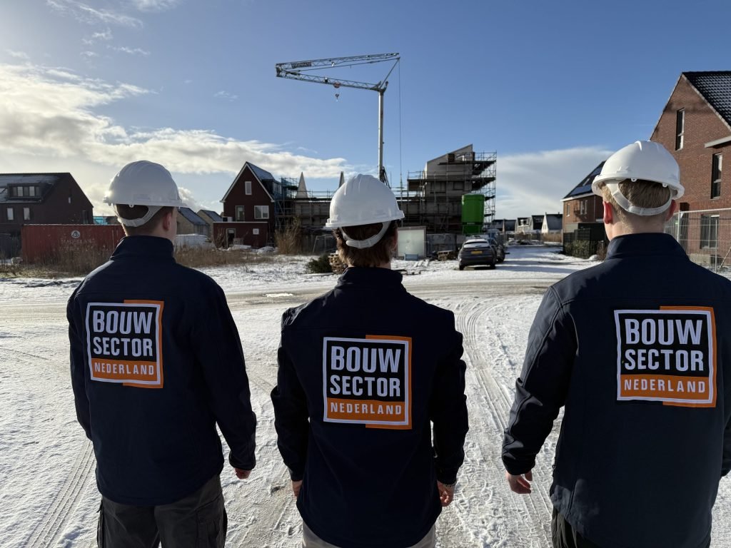 Drie vakmensen van Bouwsector Nederland met witte helm, op de rug gezien bij huizen in aanbouw en een bouwkraan